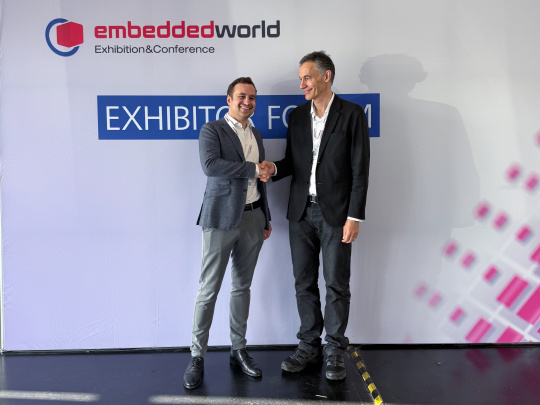 Embedded World展示会の壁の前で握手を交わす二人の代表取締役